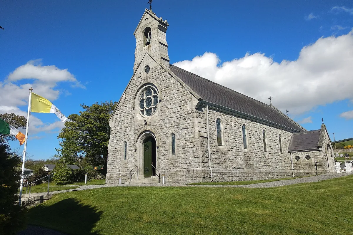 A small grey stone church with a bell tower and round window stands on a green lawn under a blue sky with white clouds.