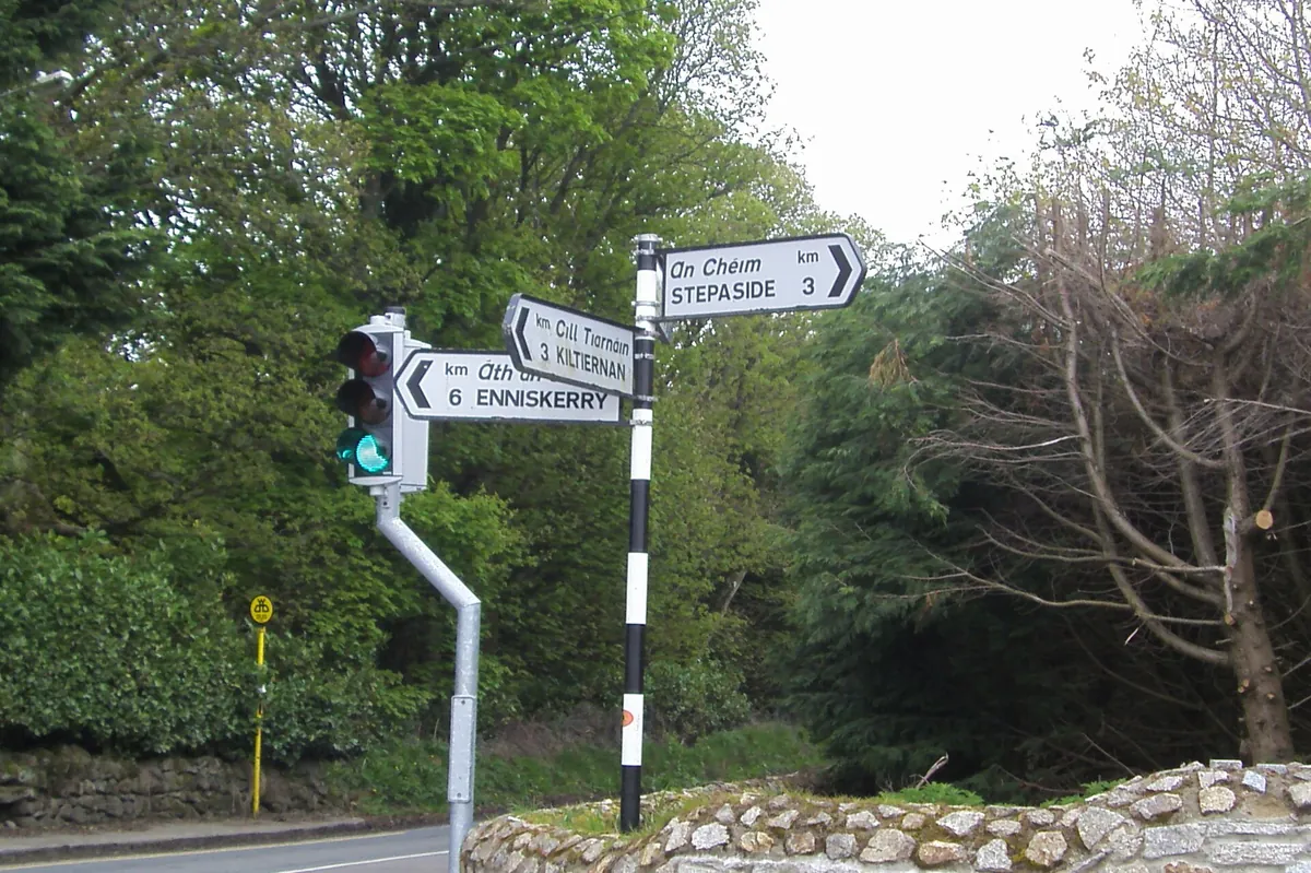 Road signs on a black-and-white post at a country crossroads pointing to Stepaside, Kilternan and Enniskerry, with a stone wall and trees behind