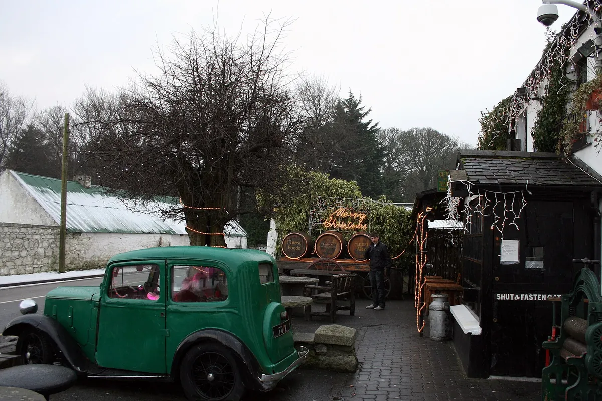 A vintage green car and wooden barrels stand on a cobbled courtyard beside the dark timber porch of a pub, with bare trees and a white outbuilding behind