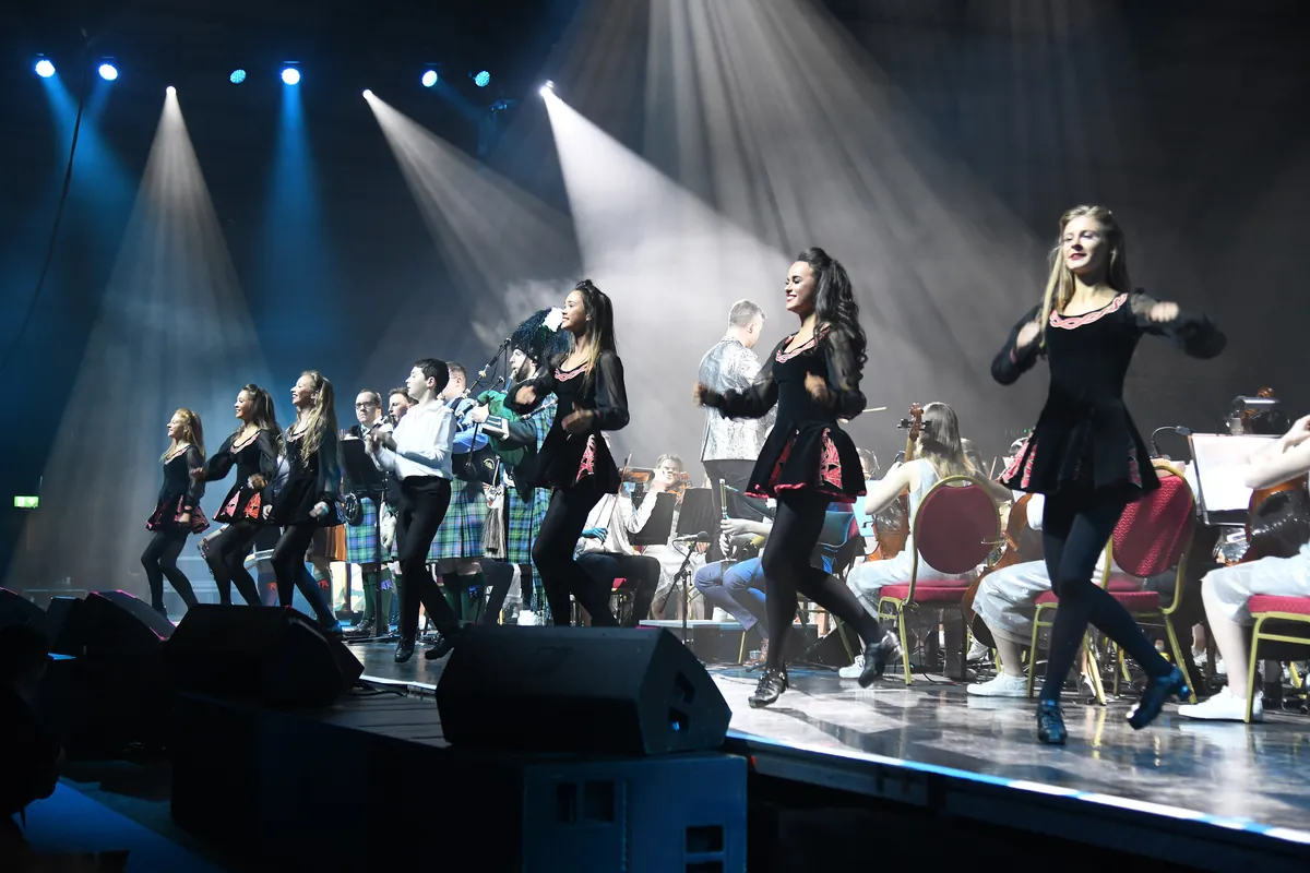 Irish dancers in black dresses and kilts leap across a lit stage in front of a line of seated musicians under shafts of blue and white light.