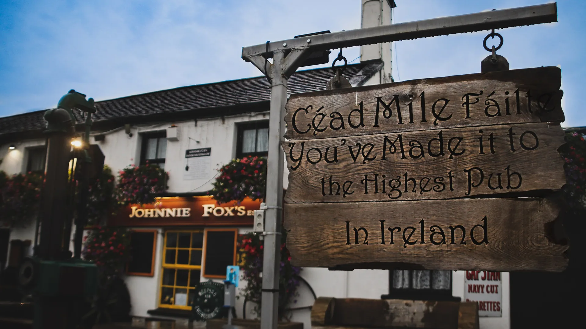 A carved wooden sign hangs from an iron post at dusk, reading Céad Mile Fáilte, You've made it to the highest pub in Ireland, in front of the white pub with its glowing orange Johnnie Fox's sign